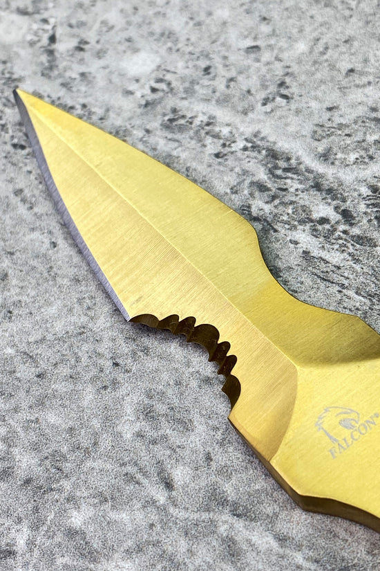 Gold finished falcon tactical knife fixed blade with partial serration and Falcon logo on stone surface