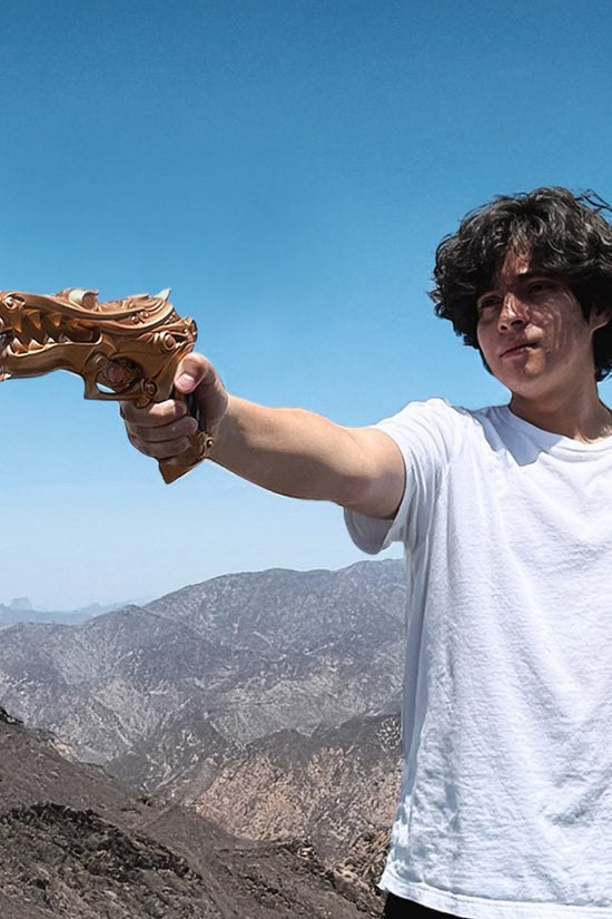 Person holding Otaku Gear foam fantasy gun prop shaped like a golden dragon pointed toward mountains and blue sky