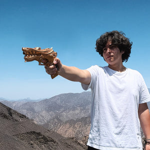 Person holding Otaku Gear foam fantasy gun prop shaped like a golden dragon pointed toward mountains and blue sky