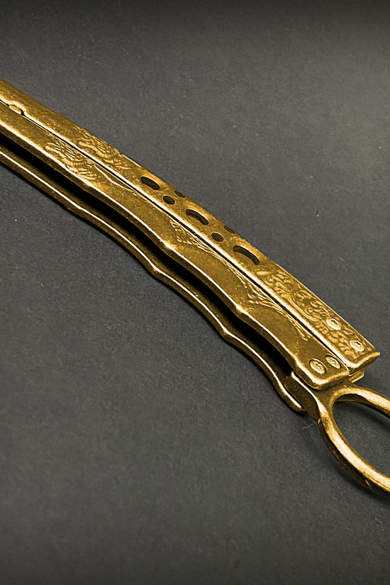 Guild Arms gold butterfly trainer knife with engraved handles on a dark surface